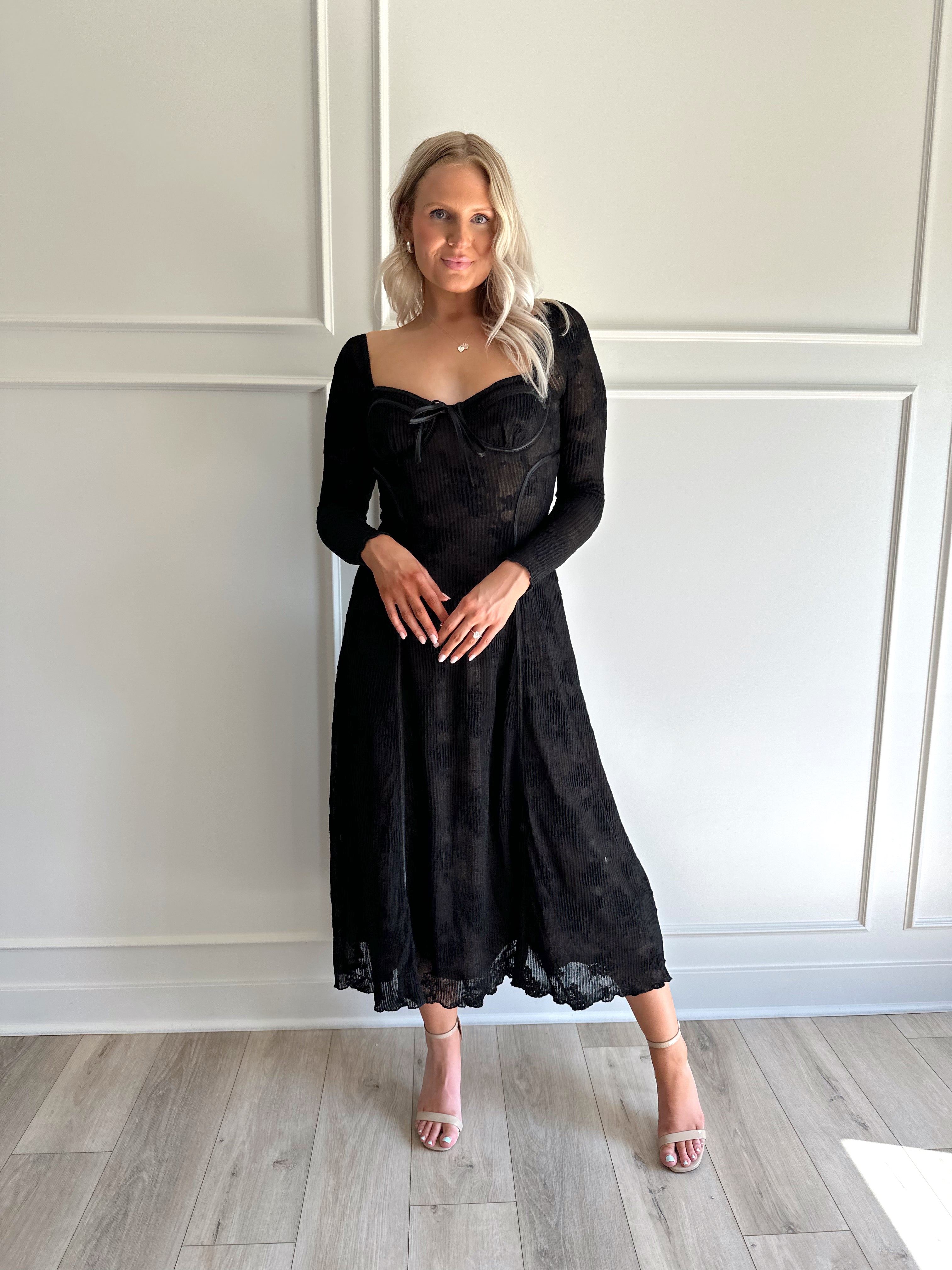 Woman wearing a black lace midi dress with long sleeves, tie detail, and flowy bottom.