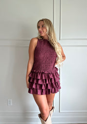 Woman wearing burgundy smocked mini dress with ruffle mini skirt and open back and tie neck.