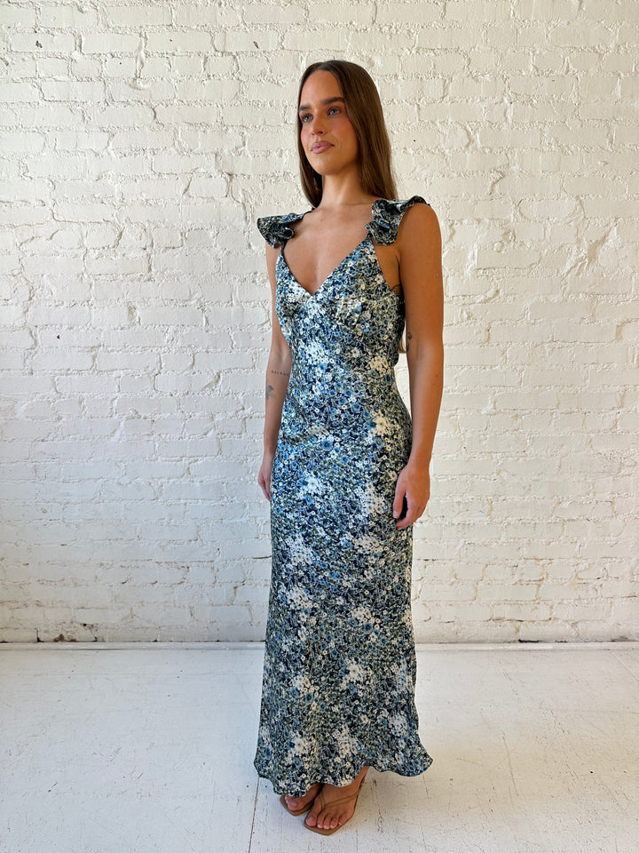 Woman wearing blue floral satin maxi dress with v-neckline, ruffle, adjustable spaghetti straps, and open back. 