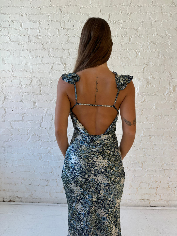 Woman wearing blue floral satin maxi dress with v-neckline, ruffle, adjustable spaghetti straps, and open back. 
