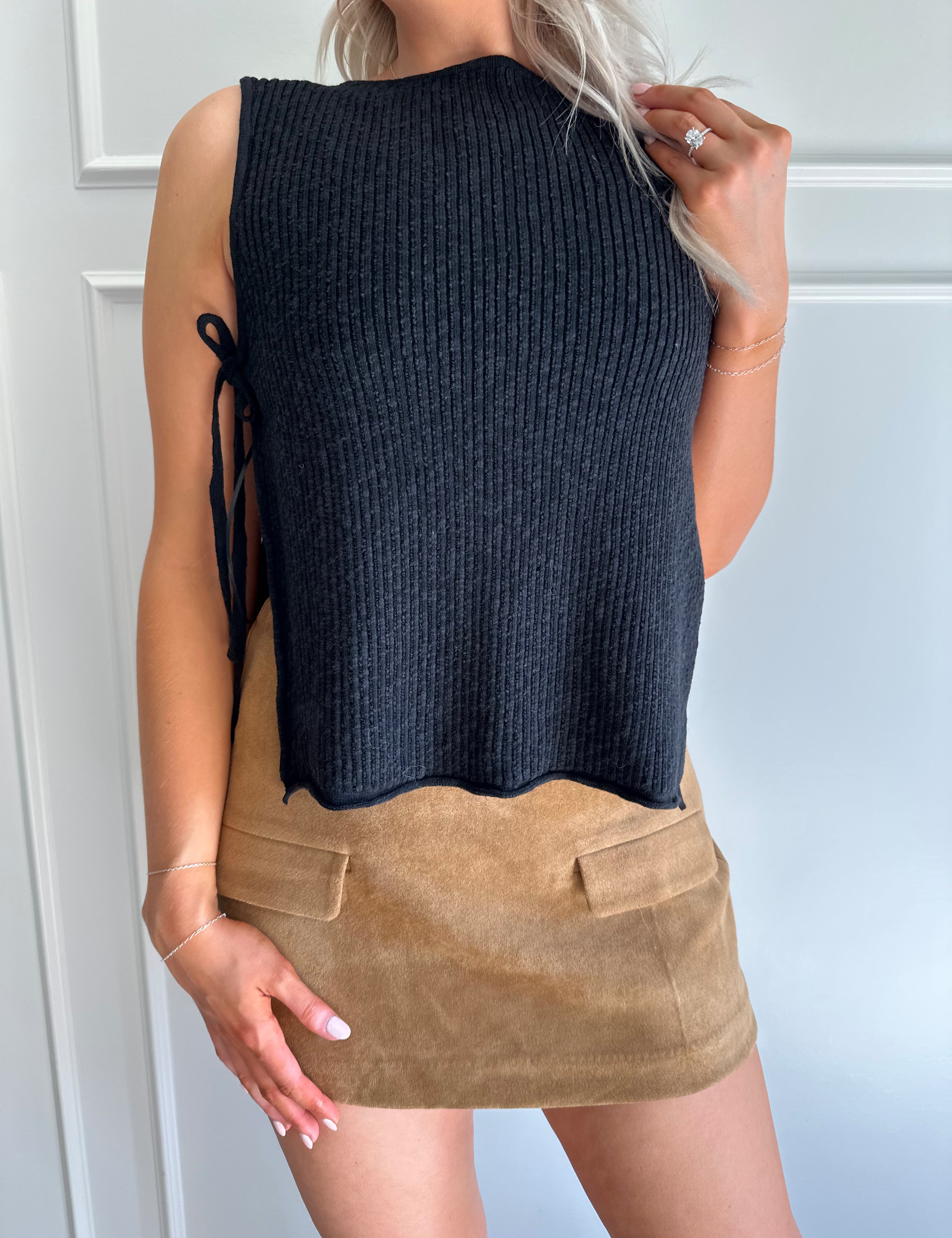 Woman wearing a black knit sleeveless sweater top with tie side detail.