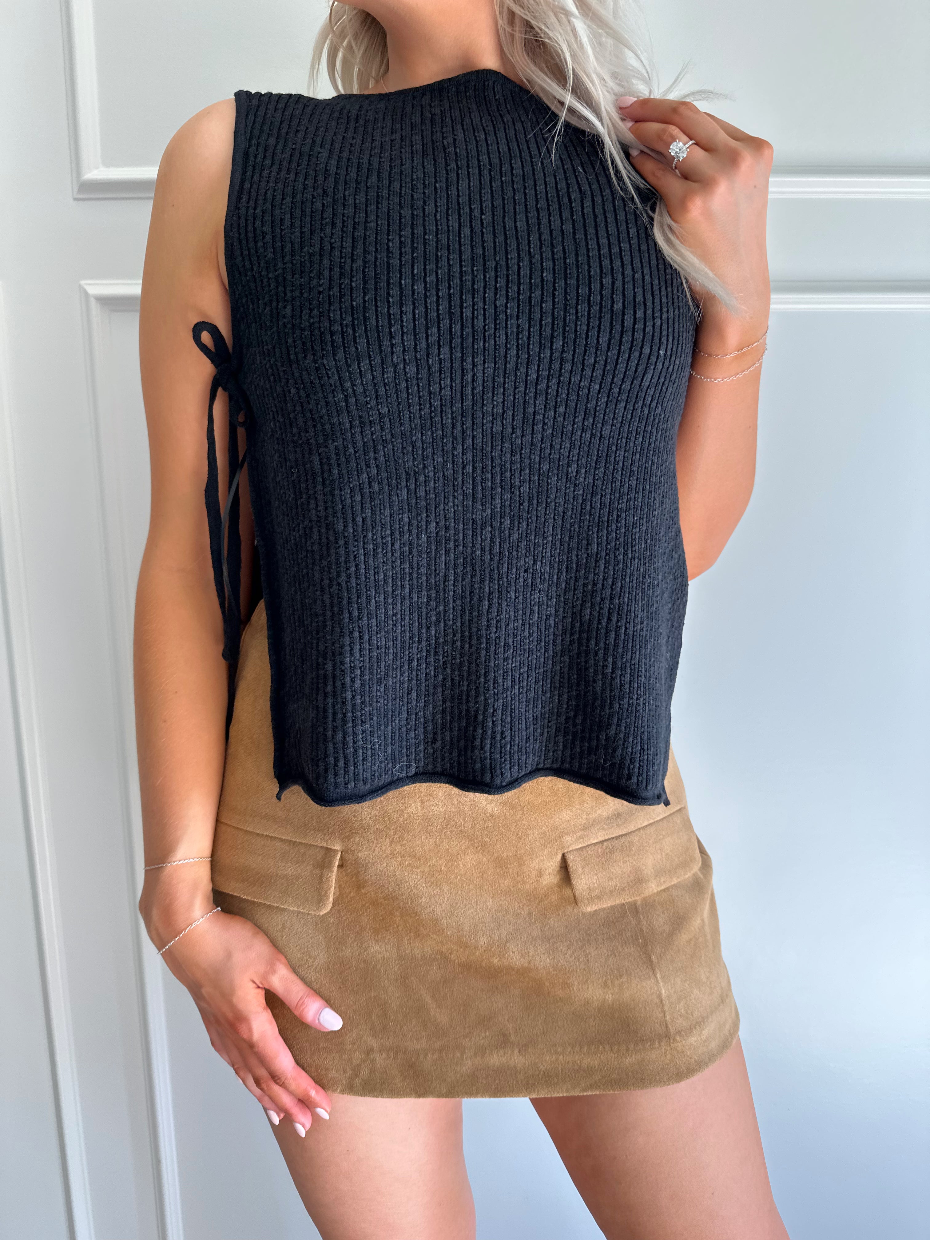 Woman wearing a black knit sleeveless sweater top with tie side detail.