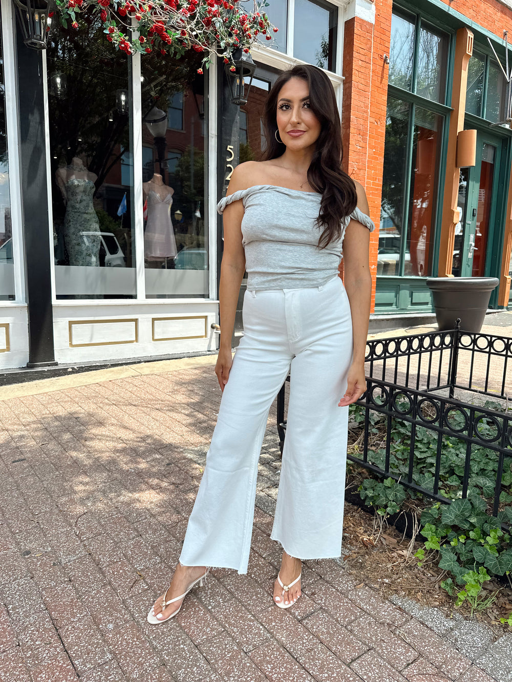 Woman wearing cream colored wide leg jean with raw hem and grey twist off the shoulder top.