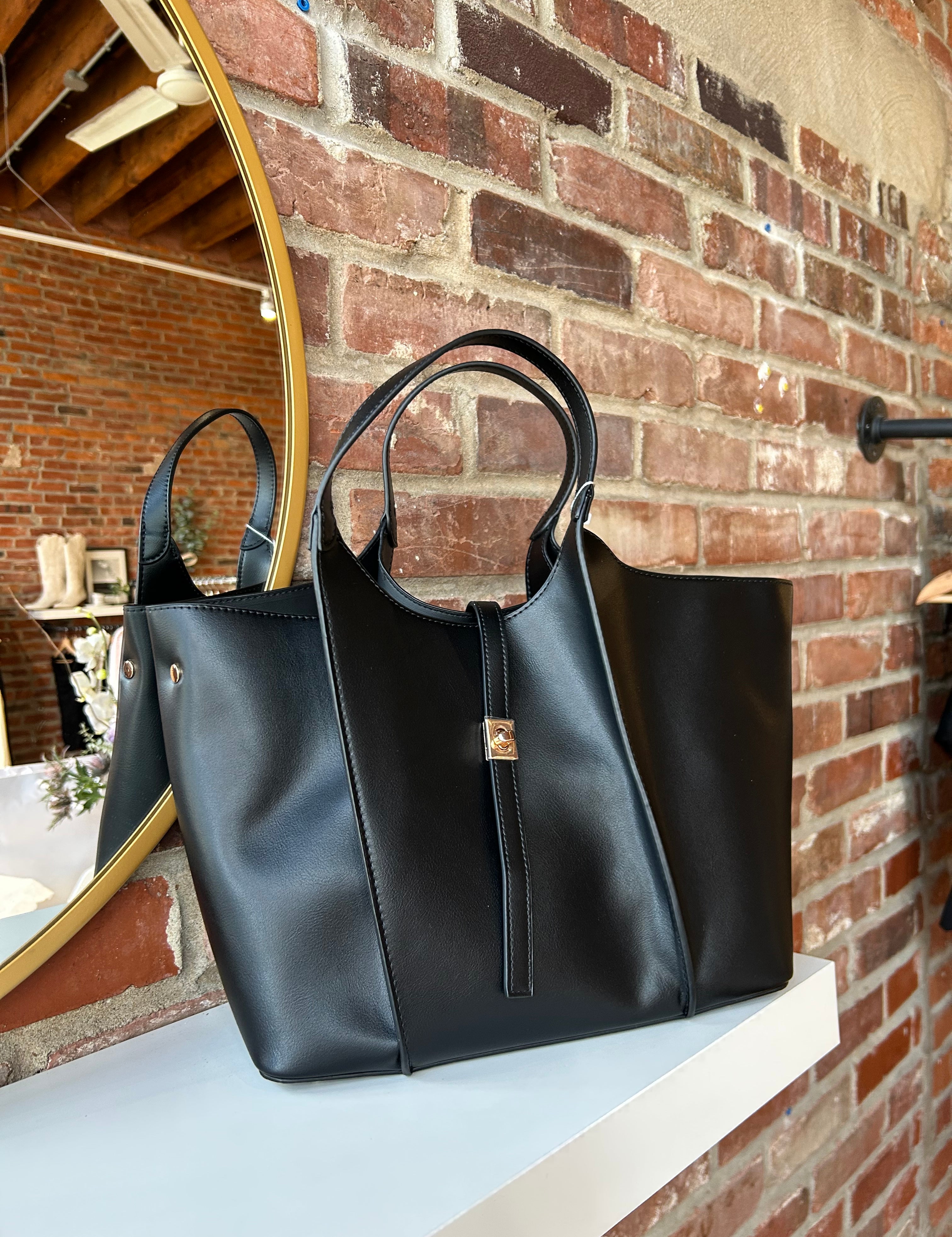 Black vegan leather tote bag with gold hardware and structured handles displayed on boutique shelf.