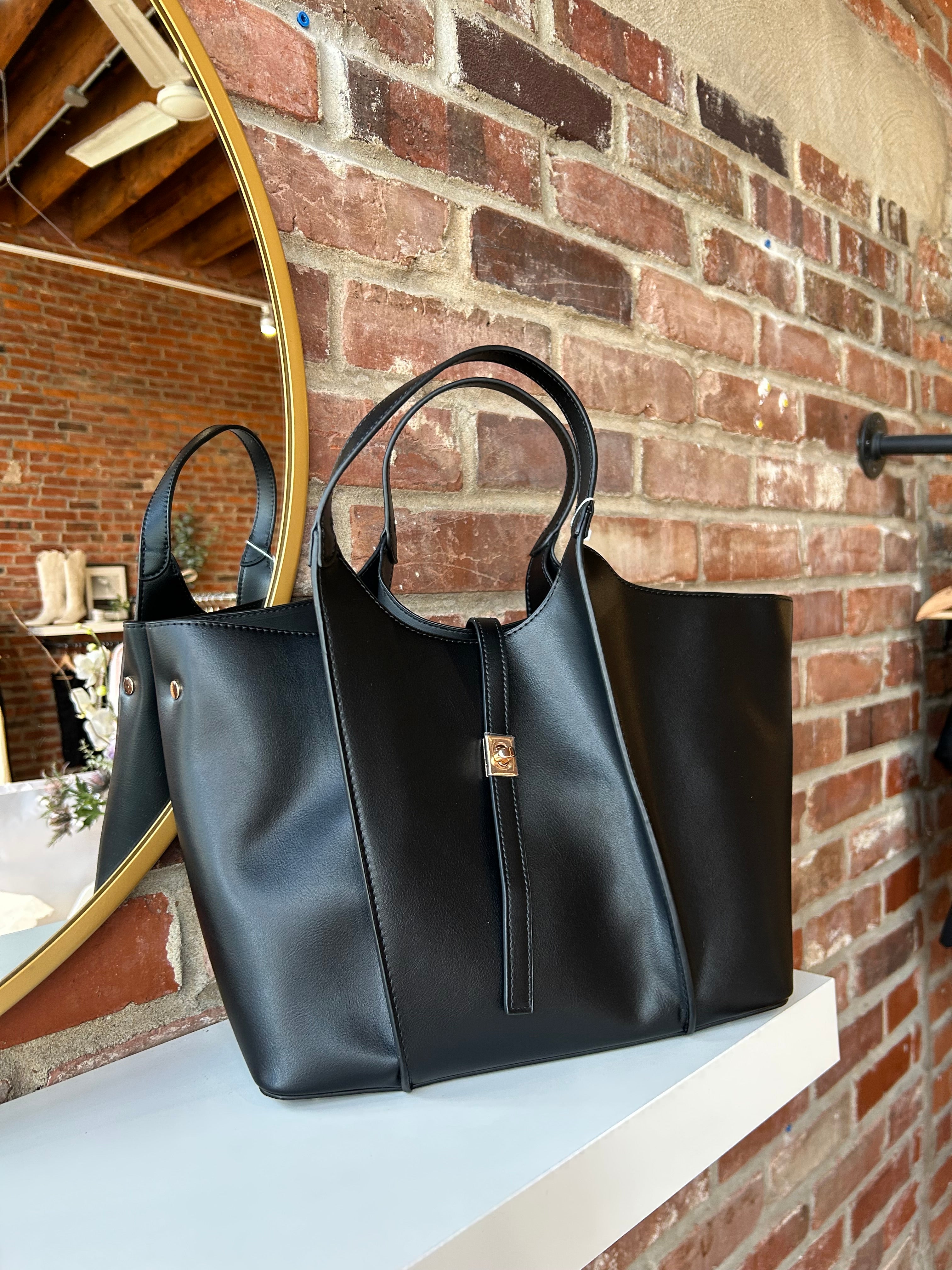 Black vegan leather tote bag with gold hardware and structured handles displayed on boutique shelf.