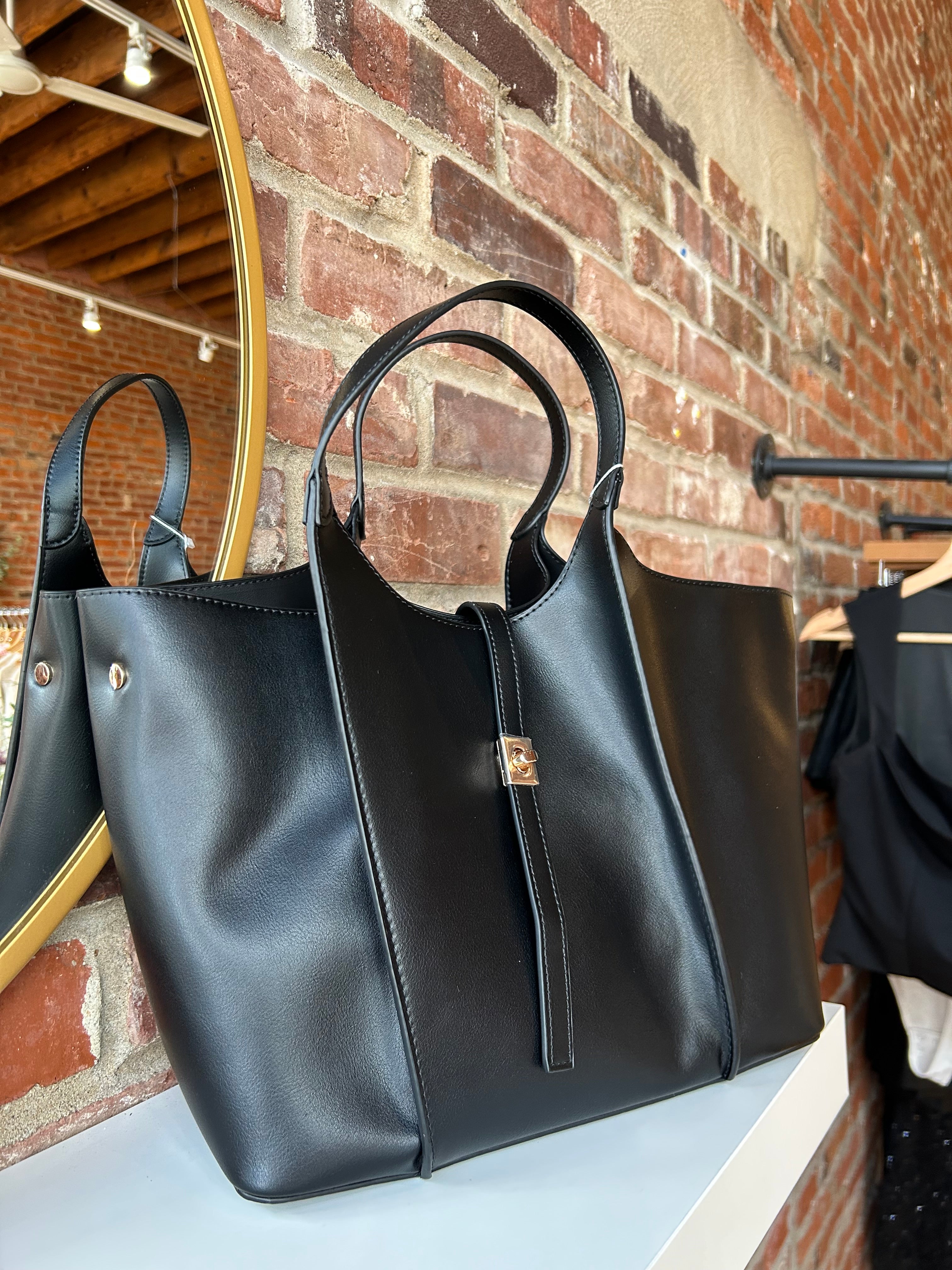 Black vegan leather tote bag with gold hardware and structured handles displayed on boutique shelf.