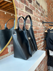 Black vegan leather tote bag with gold hardware and structured handles displayed on boutique shelf.