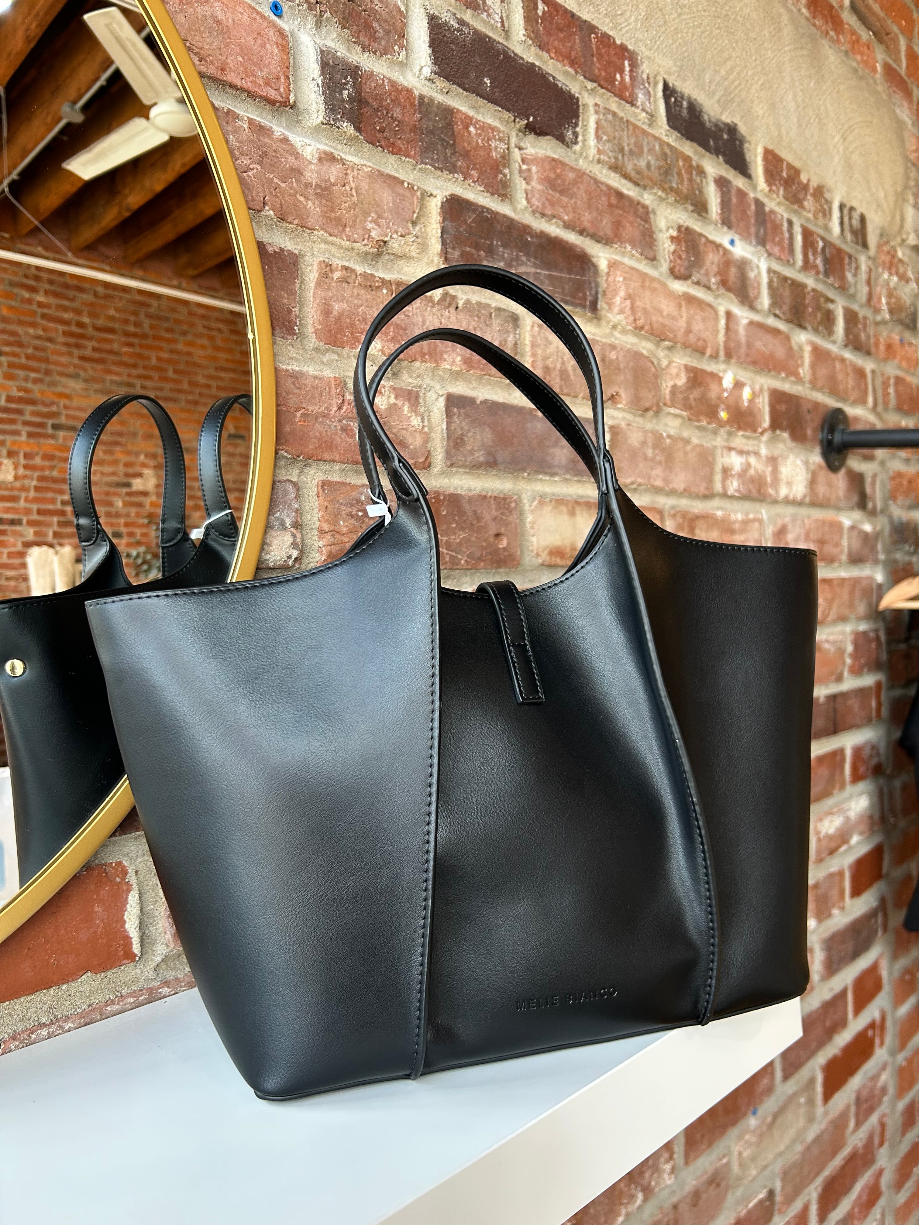 Black vegan leather tote bag with gold hardware and structured handles displayed on boutique shelf.