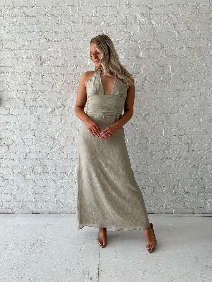 Woman wearing khaki colored maxi dress with halter neckline, and shell trim detailing. 