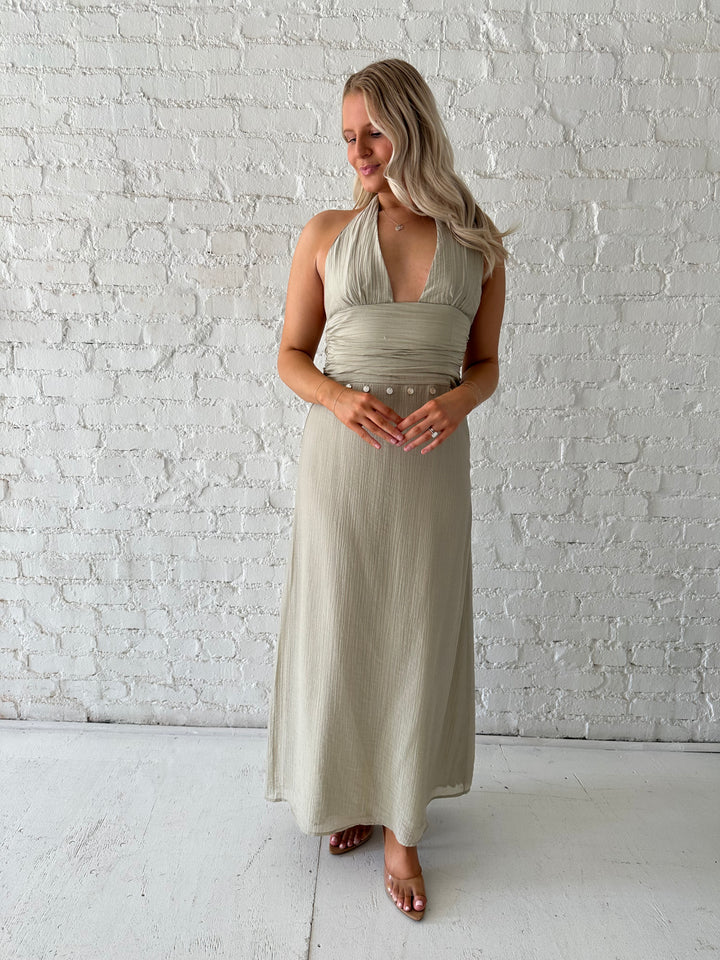 Woman wearing khaki colored maxi dress with halter neckline, and shell trim detailing. 