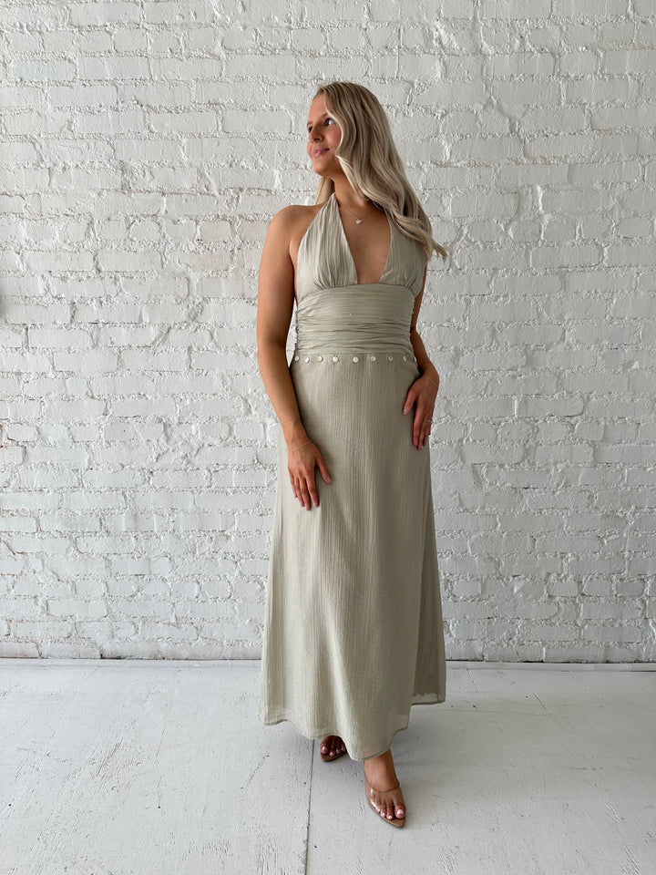 Woman wearing khaki colored maxi dress with halter neckline, and shell trim detailing. 
