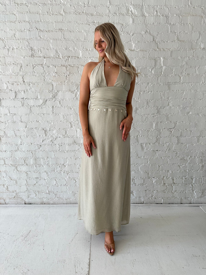 Woman wearing khaki colored maxi dress with halter neckline, and shell trim detailing. 