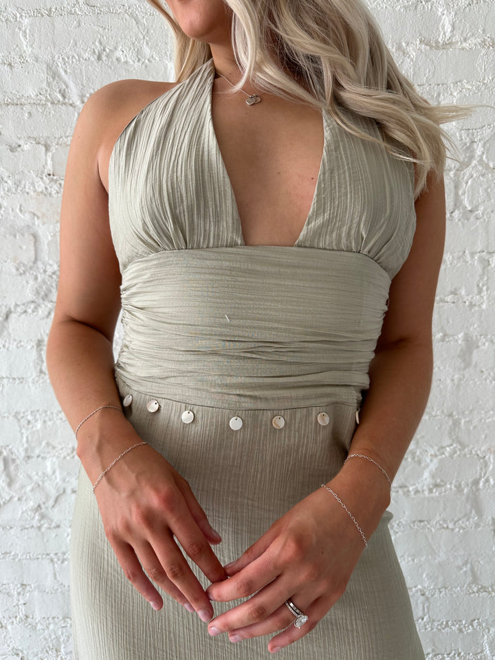 Woman wearing khaki colored maxi dress with halter neckline, and shell trim detailing. 