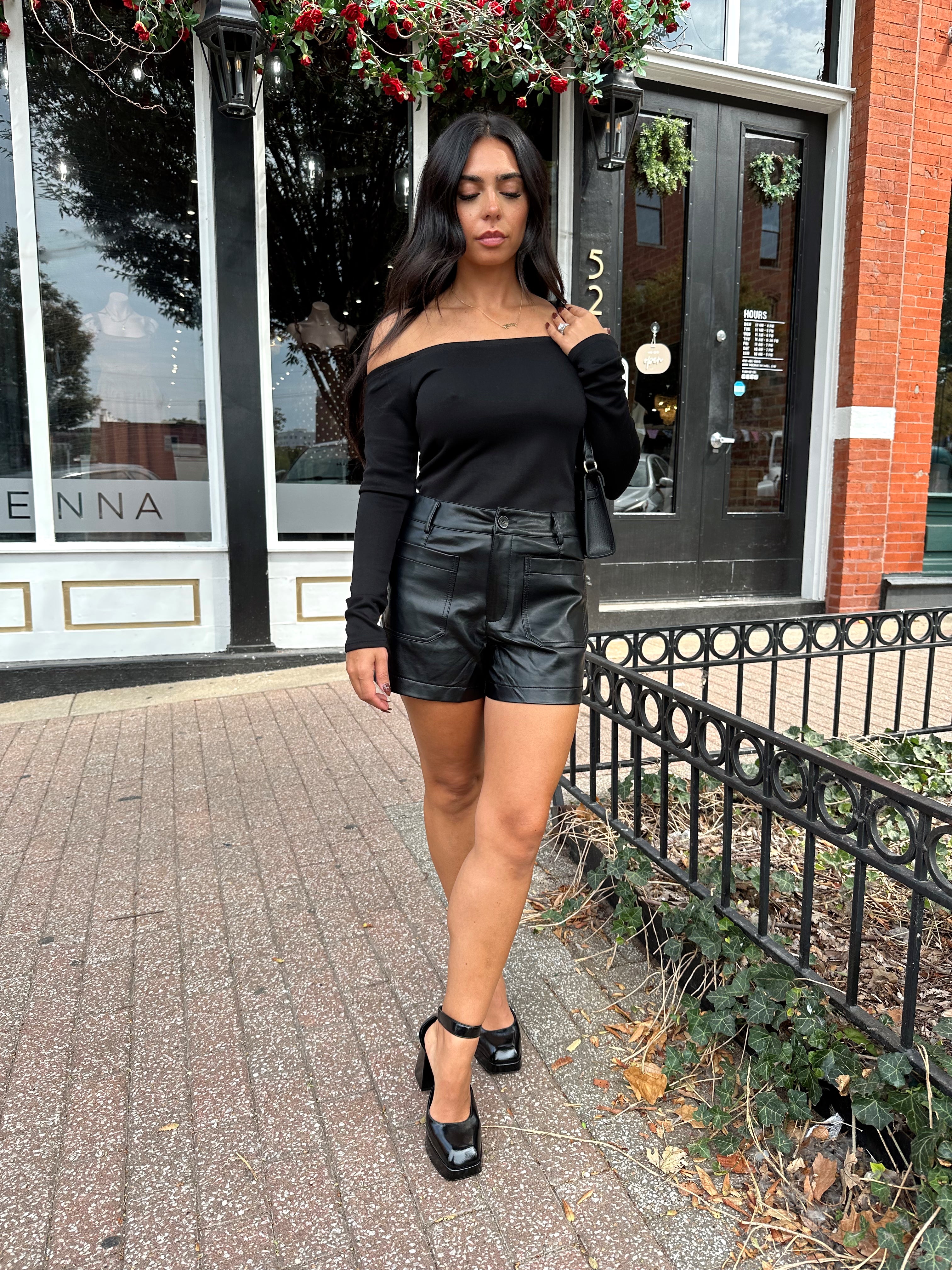 Woman wearing black off the shoulder long sleeve off the shoulder top with black leather shorts.