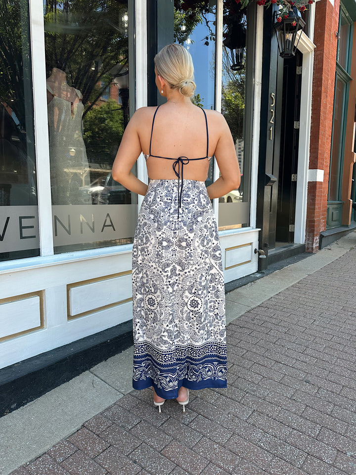 Woman wearing white and navy paisley print satin maxi dress with an open back and tie closure. 