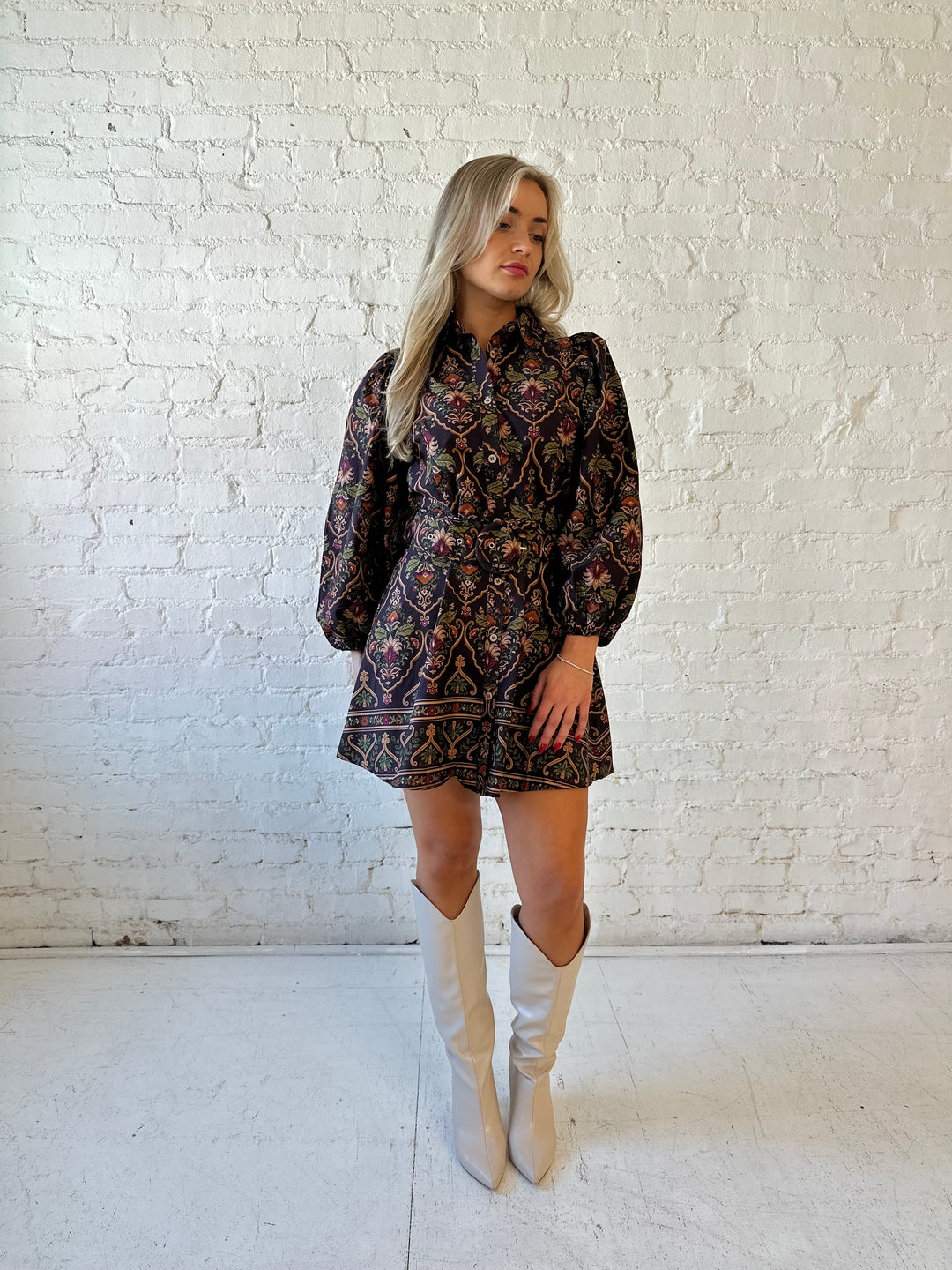 Woman wearing brown pattern long sleeve mini dress with belted waist, and knee cream boots.