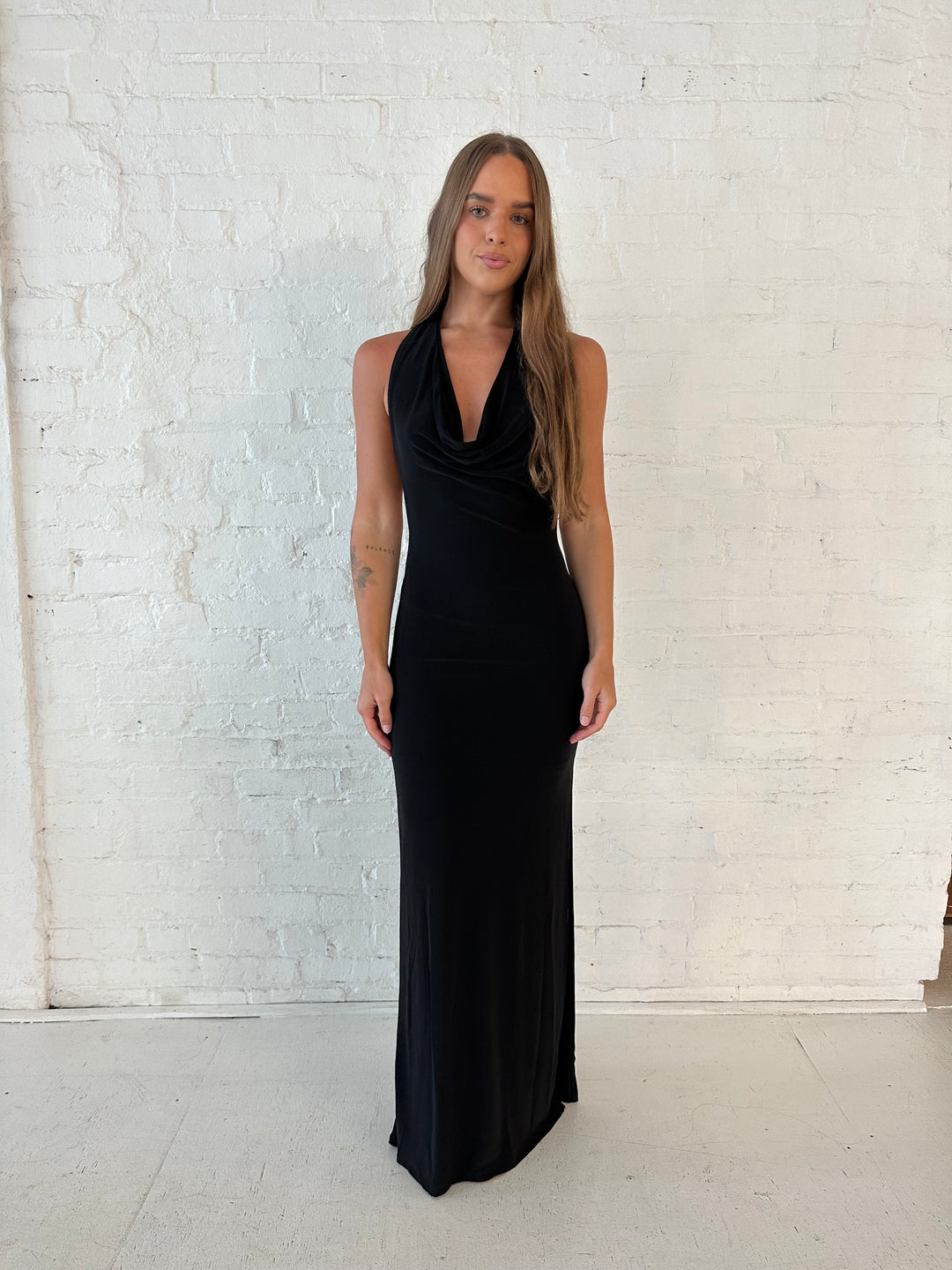 Woman wearing black maxi dress with halter cowl neckline, open back with and strap support.