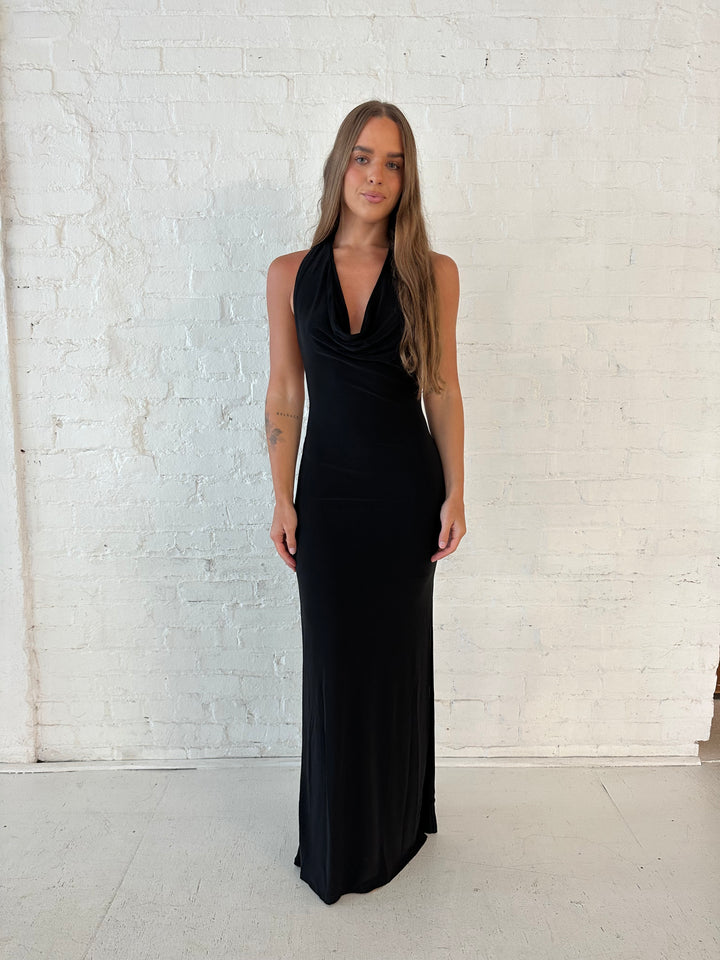 Woman wearing black maxi dress with halter cowl neckline, open back with and strap support.