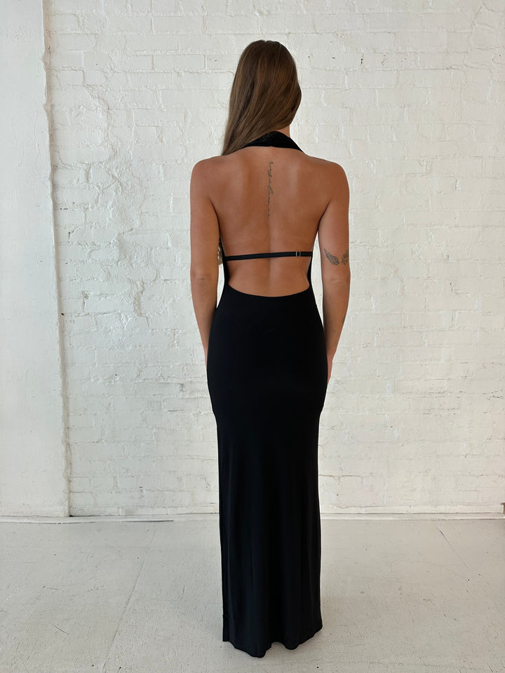 Woman wearing black maxi dress with halter cowl neckline, open back with and strap support.