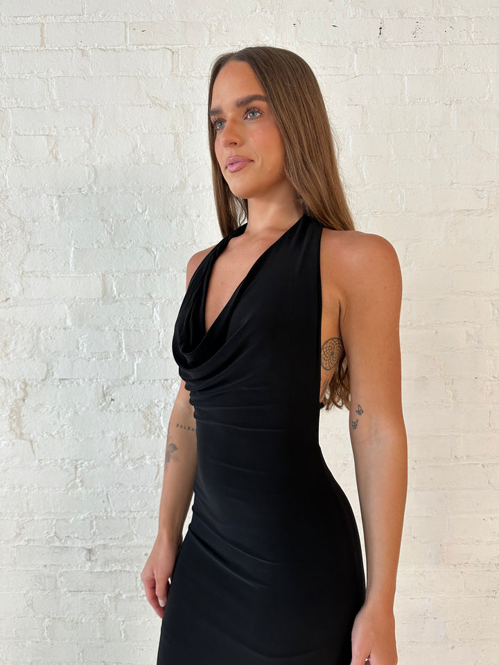 Woman wearing black maxi dress with halter cowl neckline, open back with and strap support.