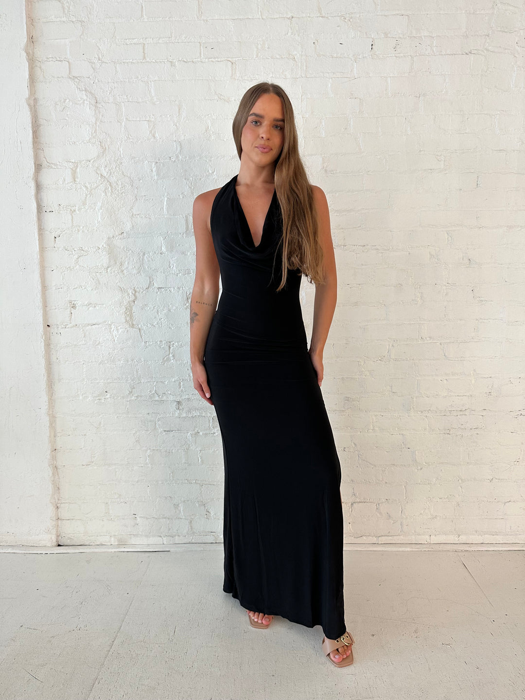 Woman wearing black maxi dress with halter cowl neckline, open back with and strap support.