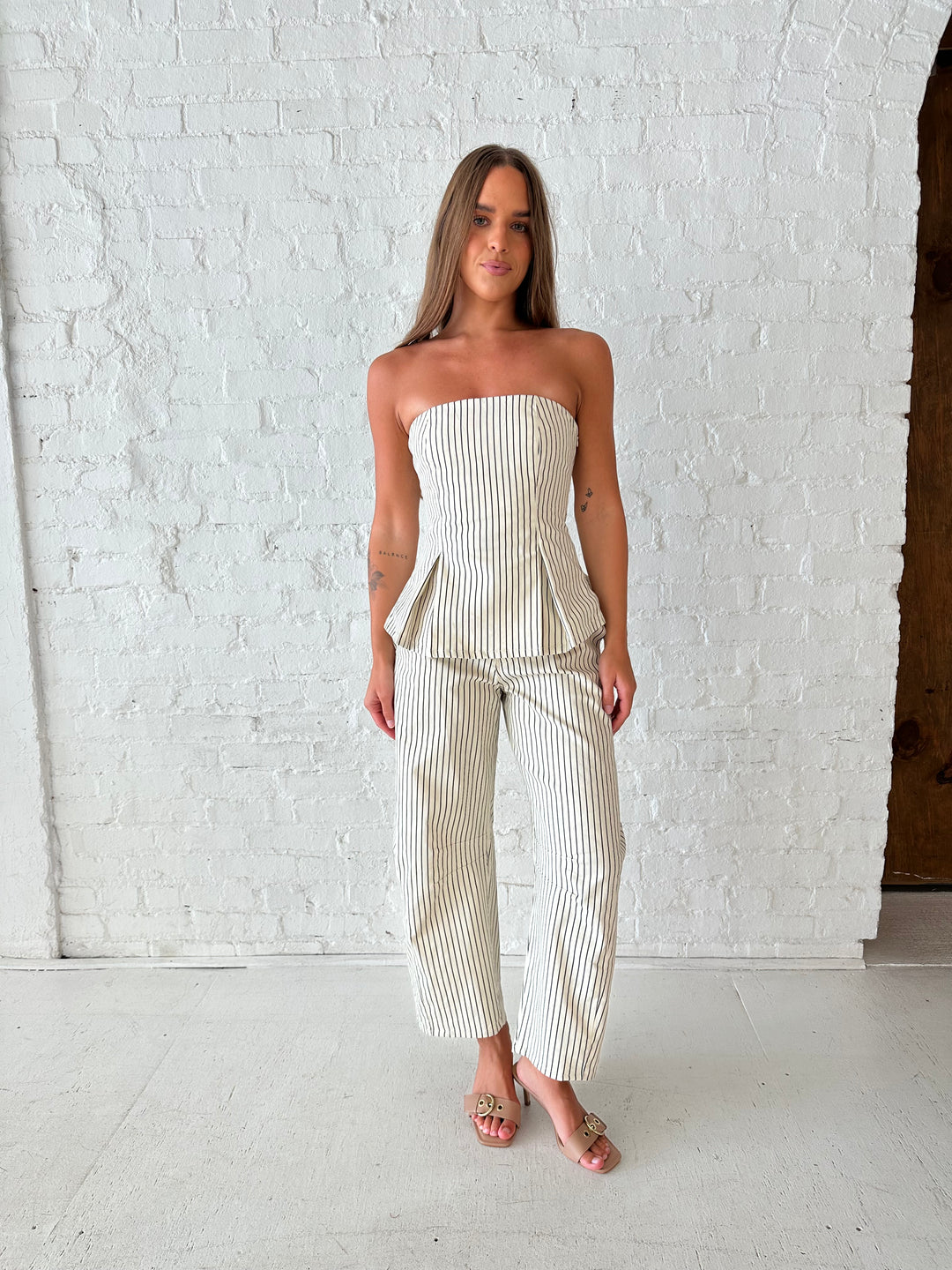 Woman wearing neutral striped two piece set with strapless peplum top, and barrel pant. 
