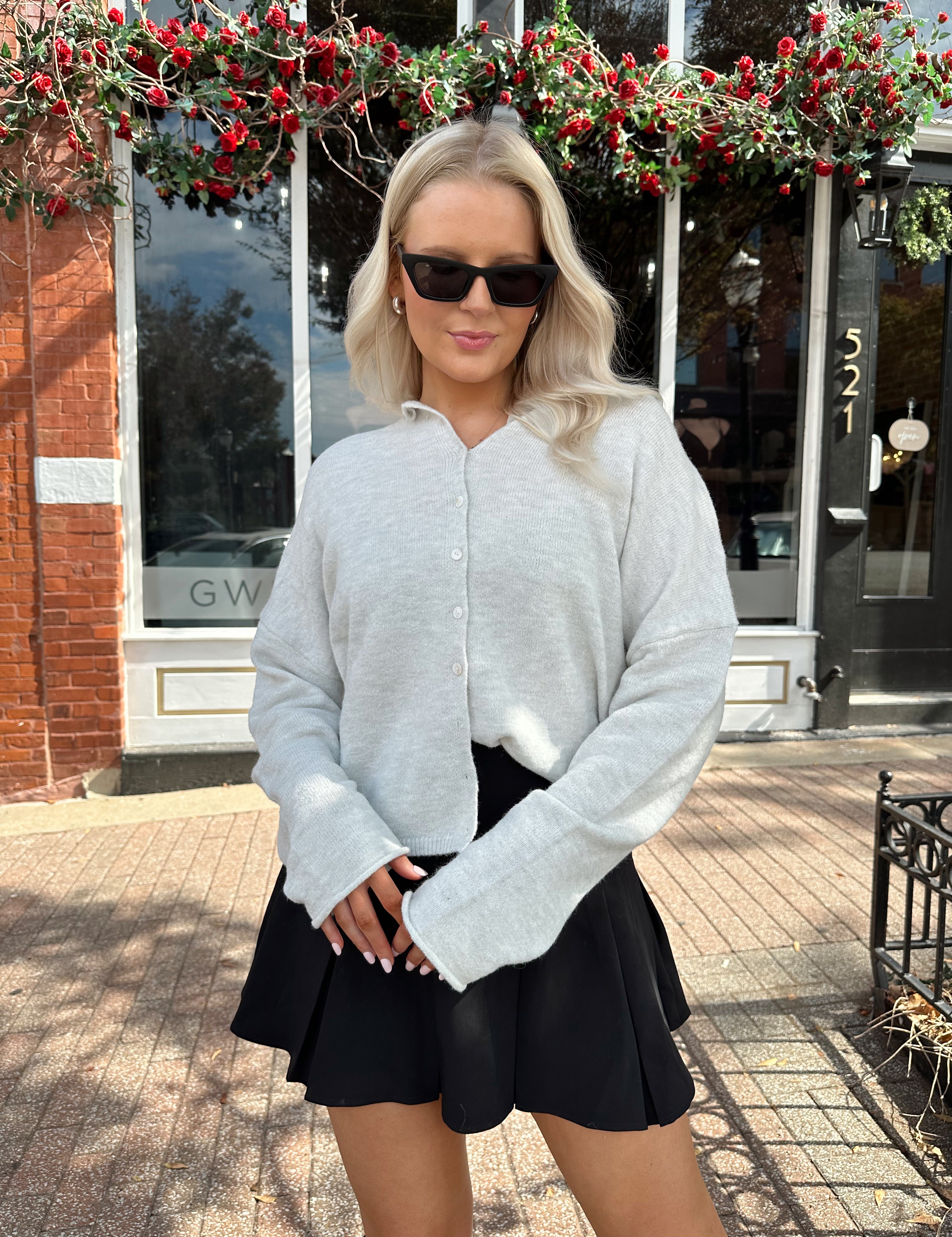 Woman wearing light grey knit button down cardigan with black pleated mini skirt. 