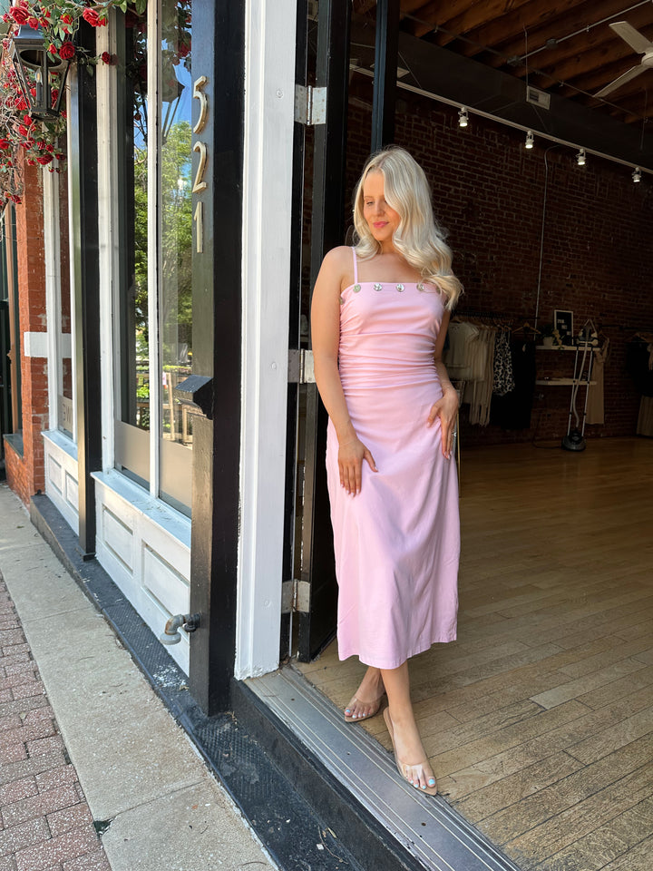 Woman wearing light pink midi dress with ruched sides, adjustable straps, and shell button detailing. 