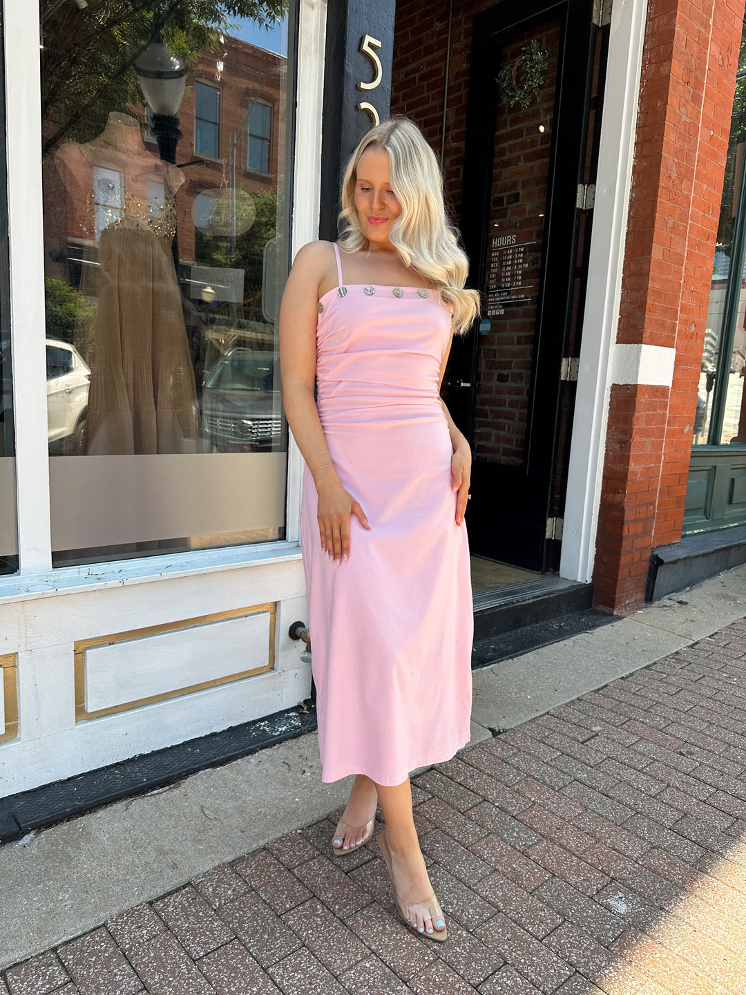 Woman wearing light pink midi dress with ruched sides, adjustable straps, and shell button detailing. 