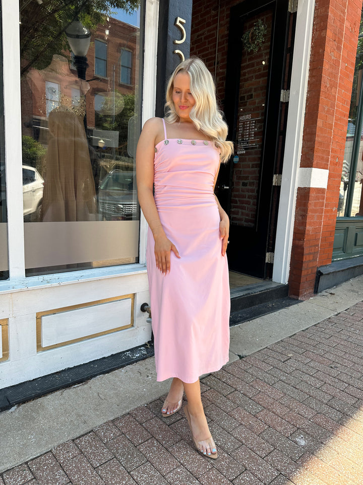 Woman wearing light pink midi dress with ruched sides, adjustable straps, and shell button detailing. 