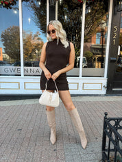 Woman wearing dark brown sleeveless sweater dress with suede knee boot.