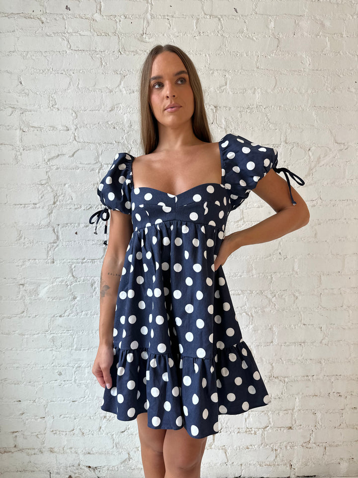 Woman wearing navy blue polka dot babydoll mini dress with sweetheart neckline, and puff sleeves.