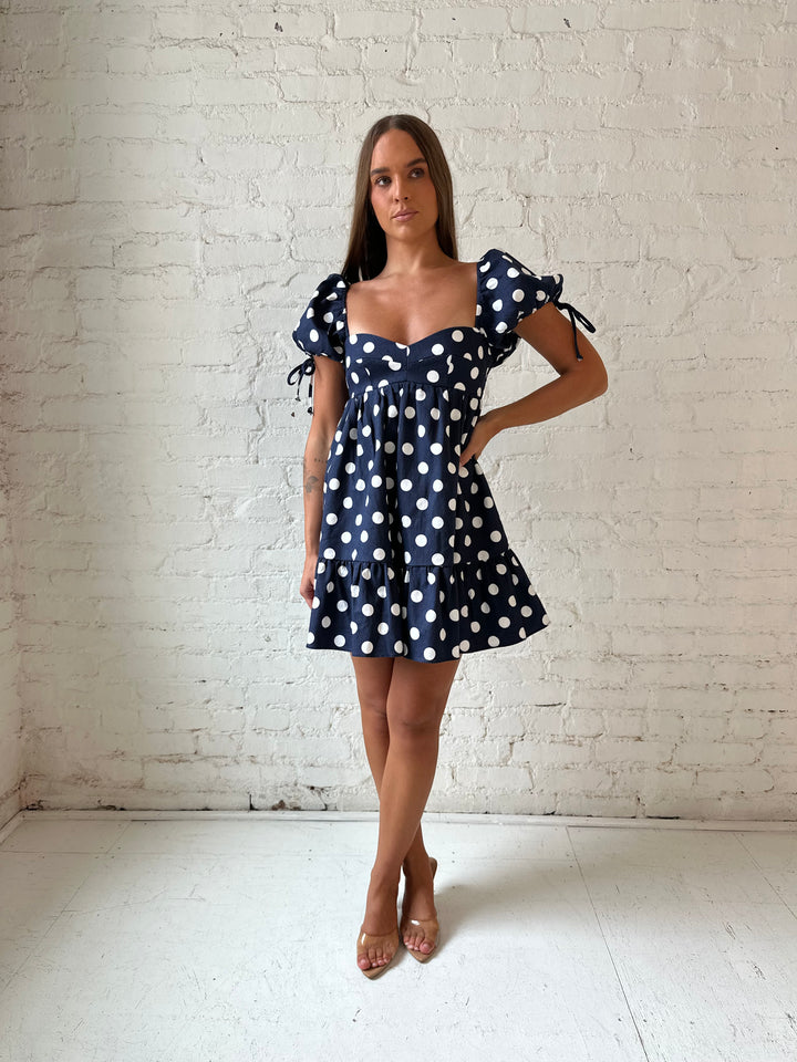 Woman wearing navy blue polka dot babydoll mini dress with sweetheart neckline, and puff sleeves.
