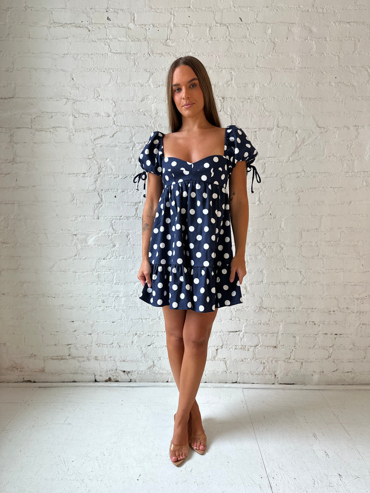 Woman wearing navy blue polka dot babydoll mini dress with sweetheart neckline, and puff sleeves.
