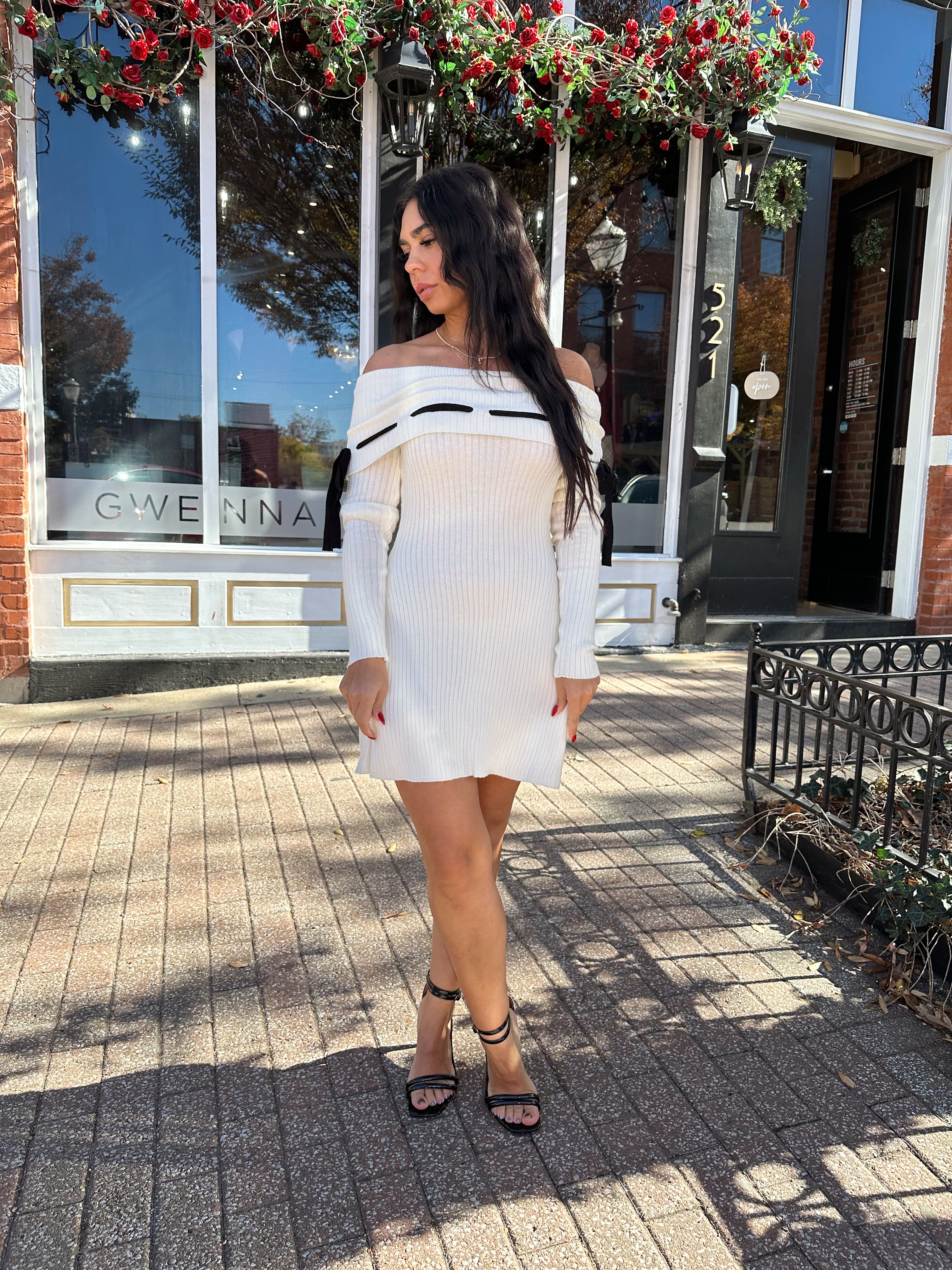 Woman wearing cream off the shoulder sweater mini dress with black tie detail. 