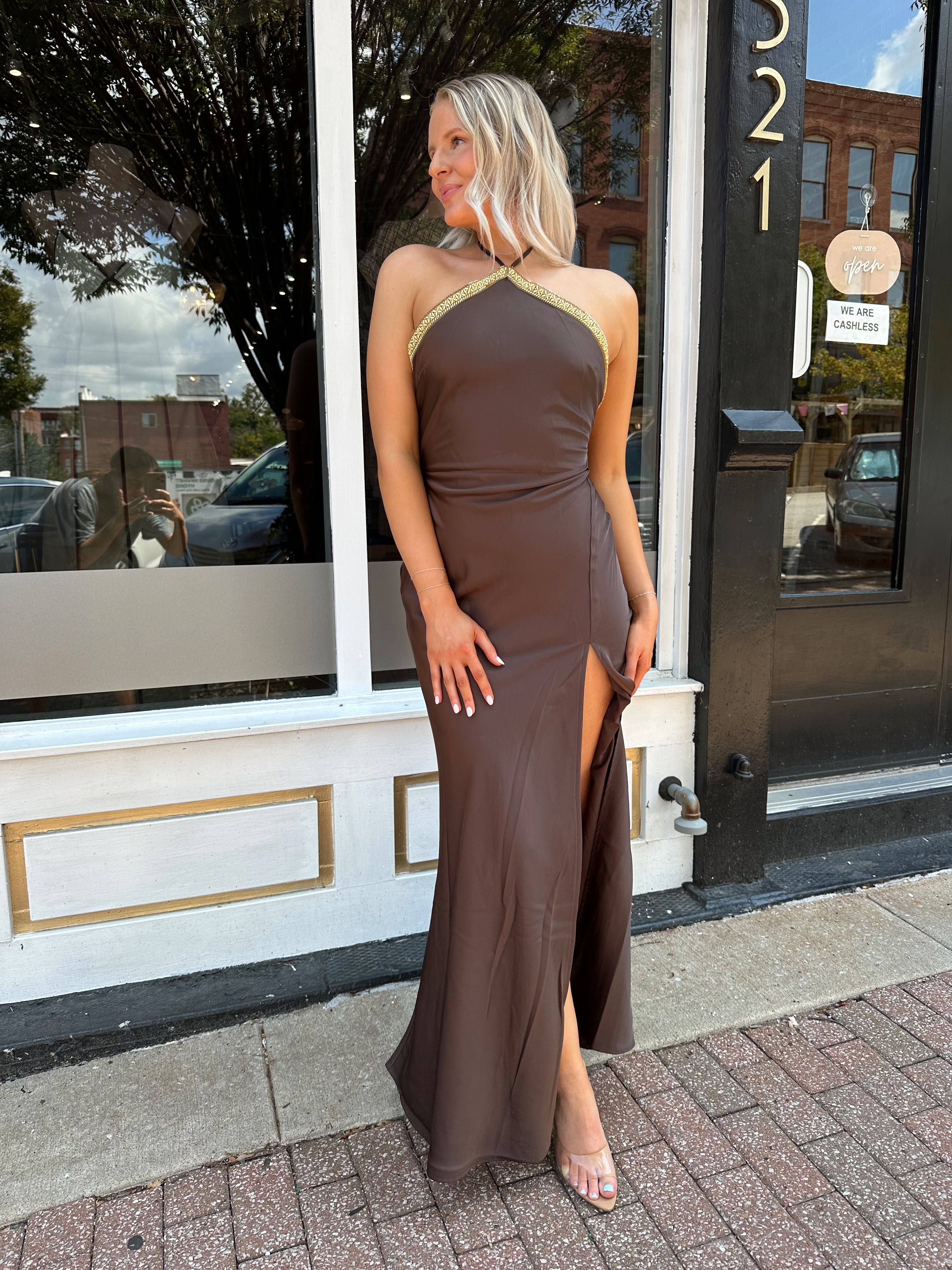 Woman wearing a dark brown satin maxi halter maxi dress with gold trim.
