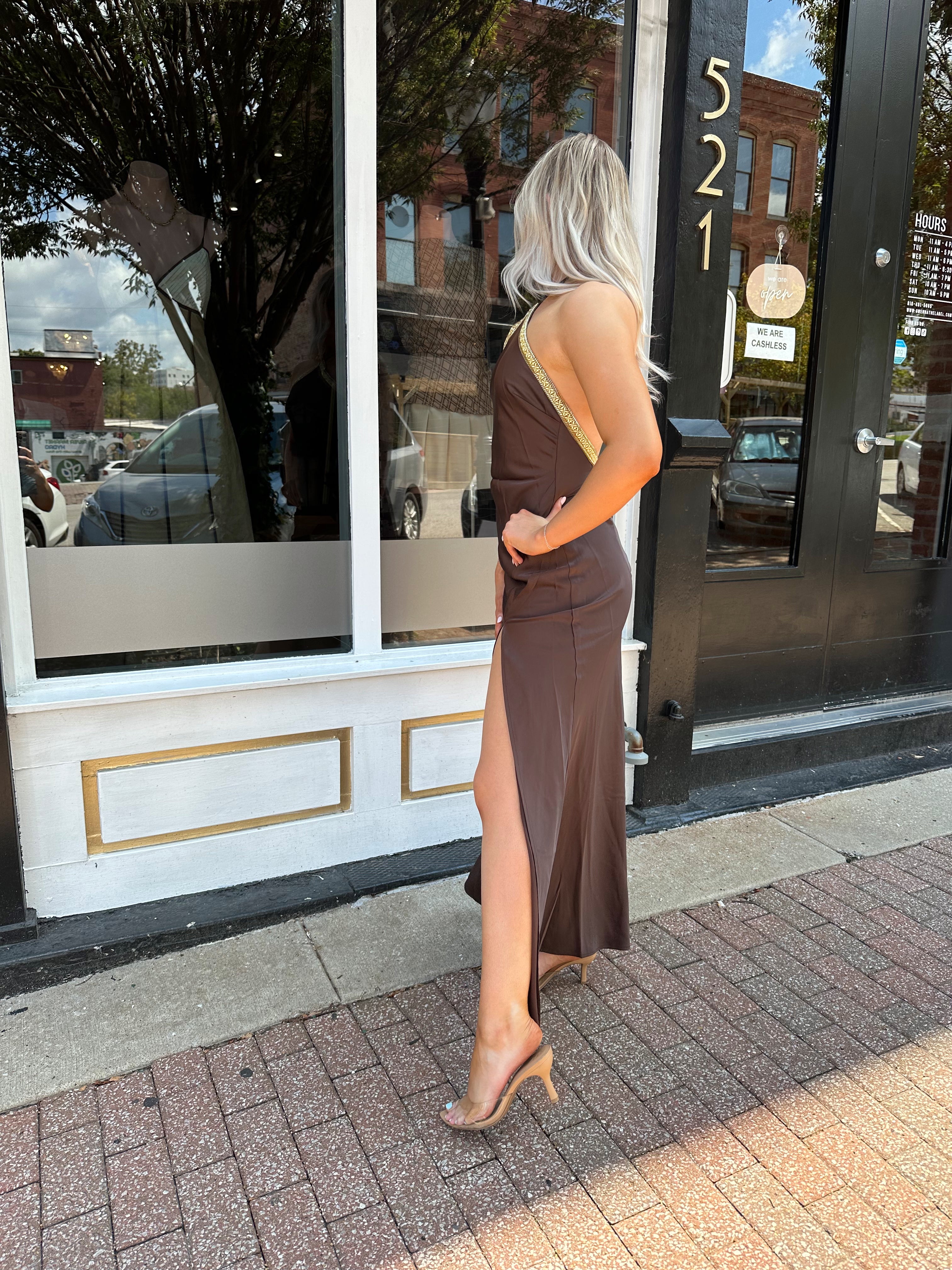 Woman wearing a dark brown satin maxi halter maxi dress with gold trim.