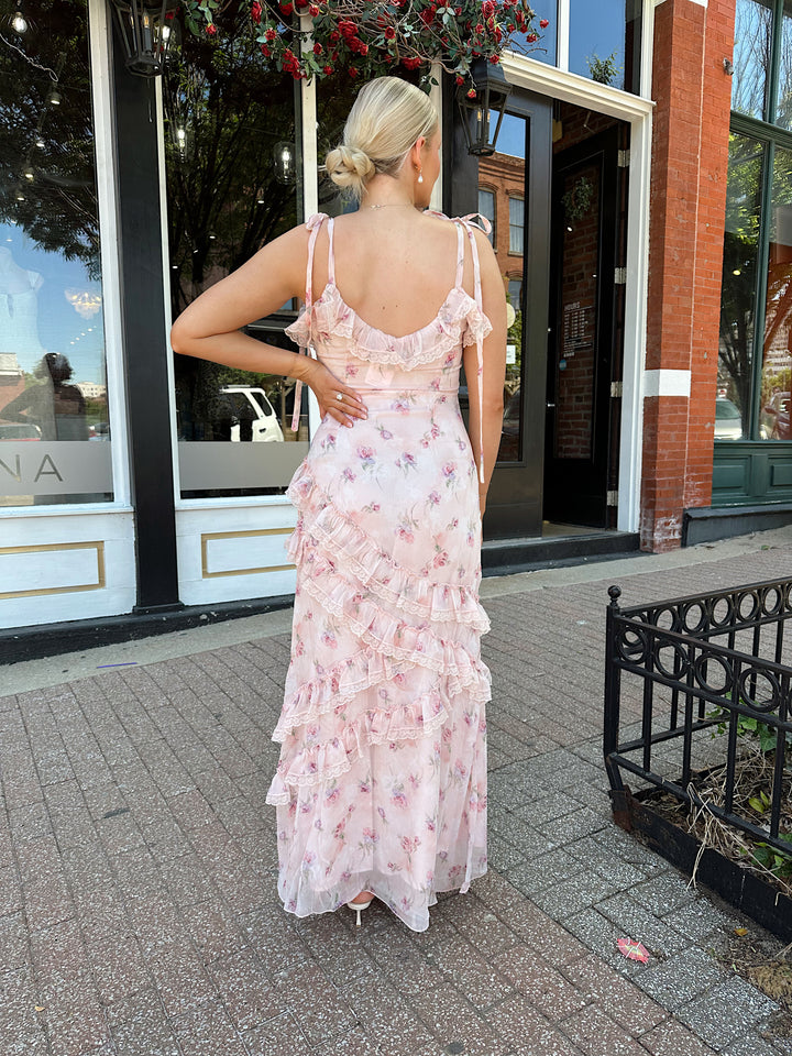 Woman wearing light pink floral ruffle tiered maxi dress with lace trim, and self tie straps.