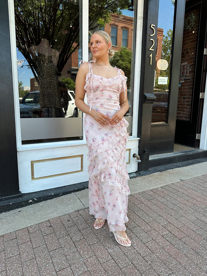 Woman wearing light pink floral ruffle tiered maxi dress with lace trim, and self tie straps.