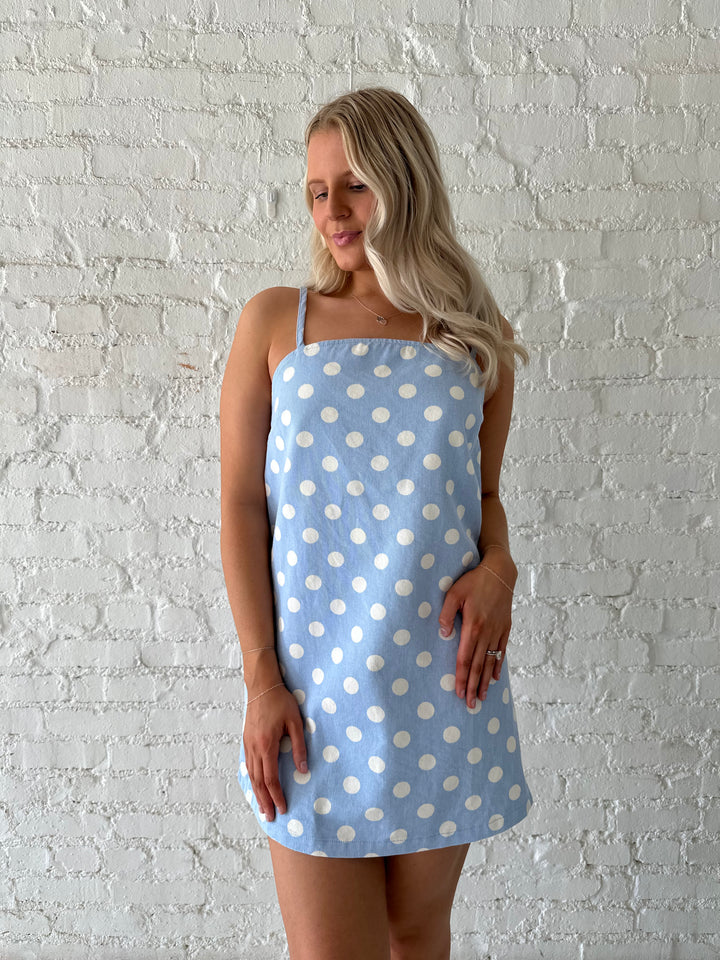Woman wearing sky blue polka dot denim mini dress with straight neckline and adjustable straps.