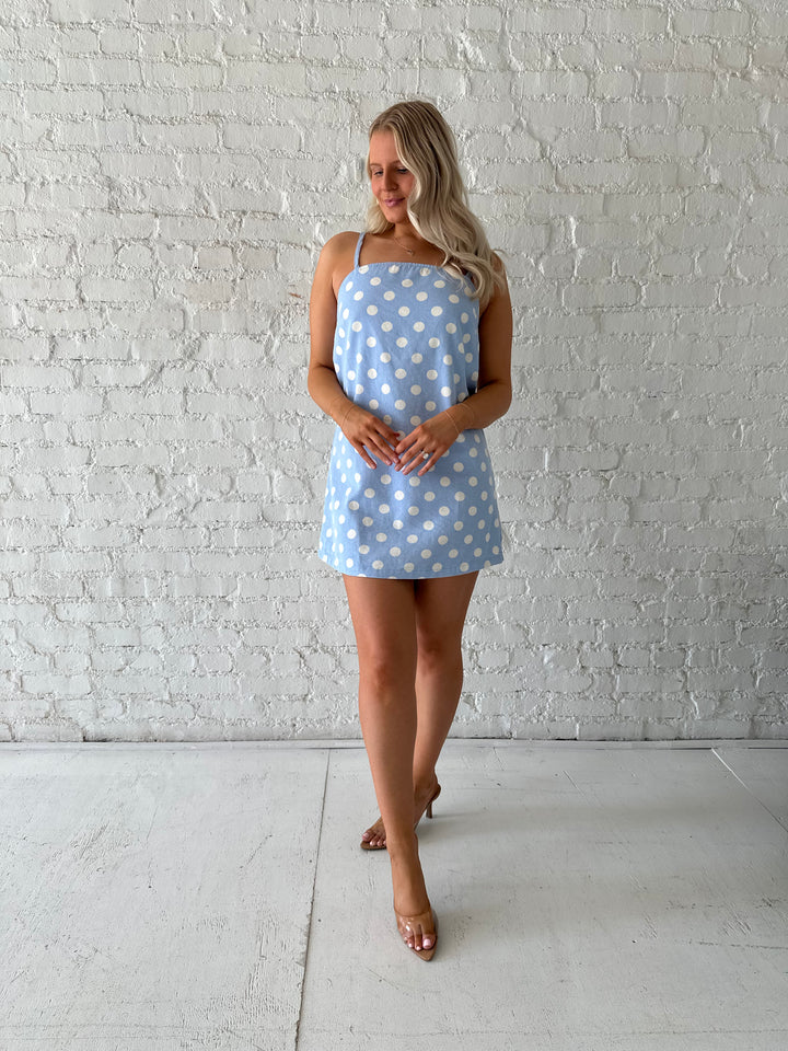 Woman wearing sky blue polka dot denim mini dress with straight neckline and adjustable straps.