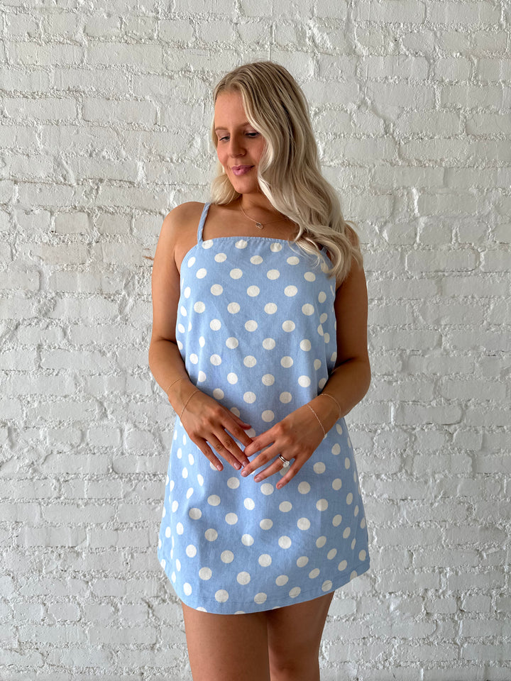Woman wearing sky blue polka dot denim mini dress with straight neckline and adjustable straps.