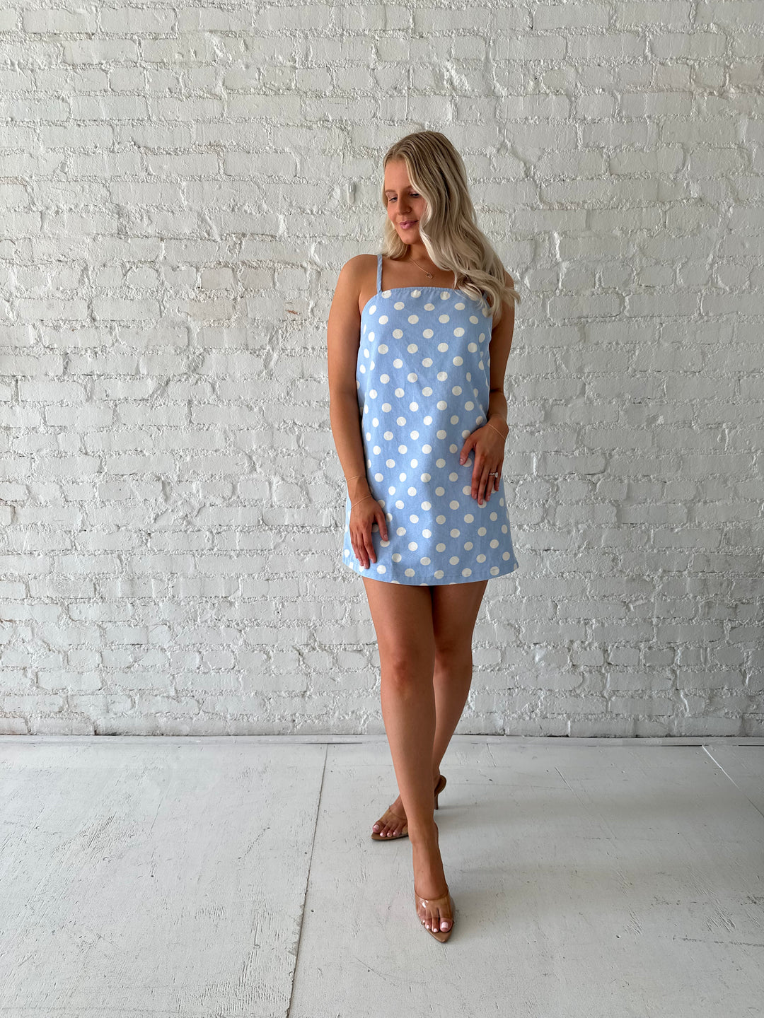 Woman wearing sky blue polka dot denim mini dress with straight neckline and adjustable straps.