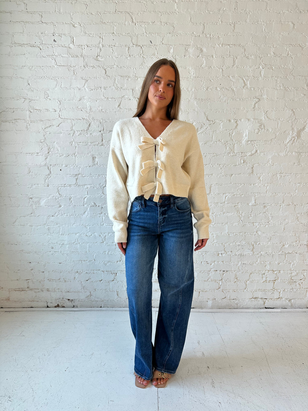 Woman wearing cream reversible bow sweater with dark wash barrel jean. 