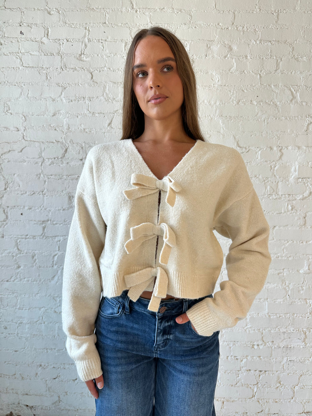 Woman wearing cream reversible bow sweater with dark wash barrel jean. 