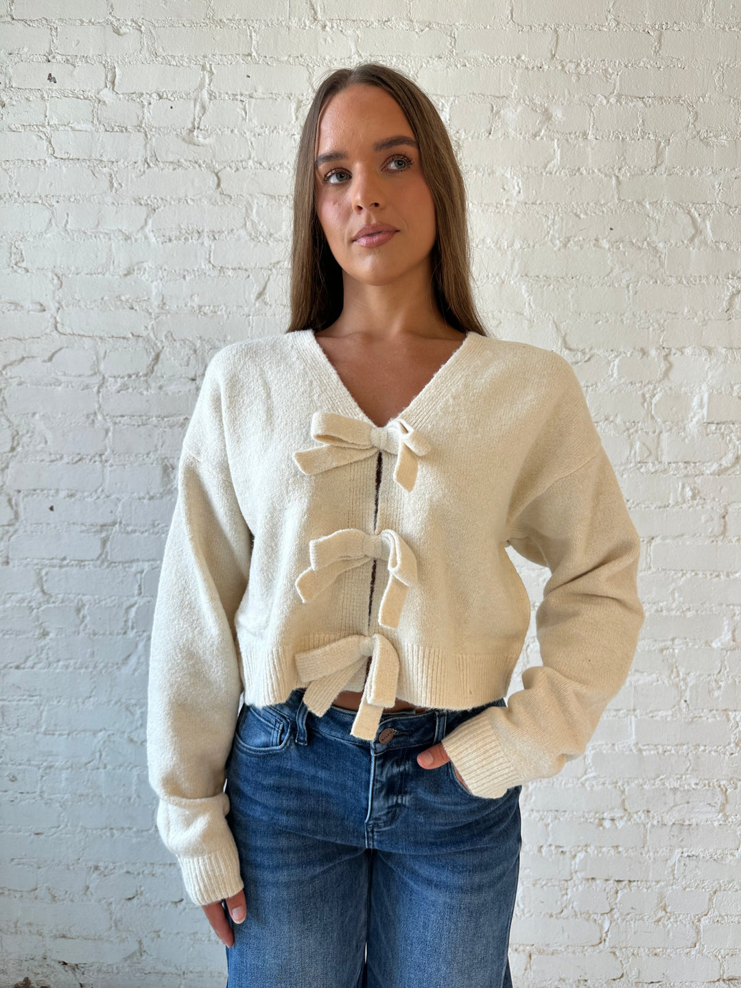 Woman wearing cream reversible bow sweater with dark wash barrel jean. 