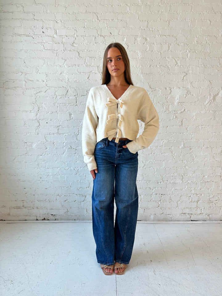 Woman wearing cream reversible bow sweater with dark wash barrel jean. 