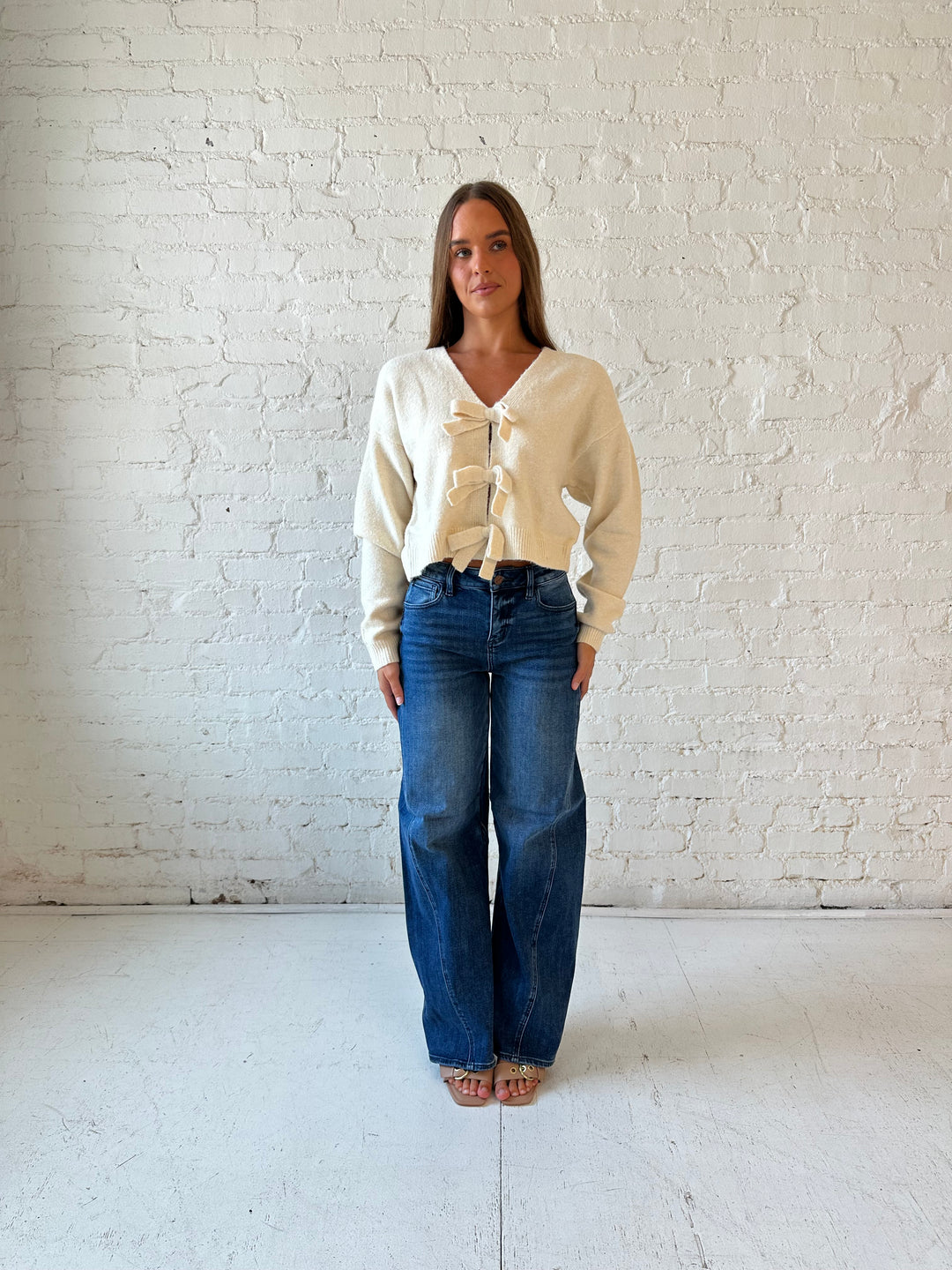 Woman wearing cream reversible bow sweater with dark wash barrel jean. 