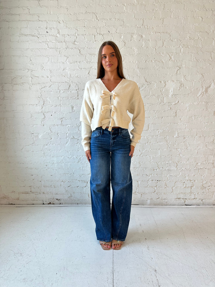 Woman wearing cream reversible bow sweater with dark wash barrel jean. 