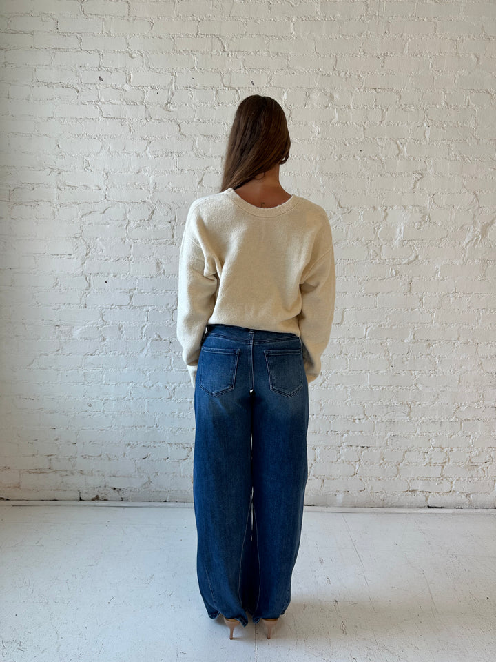 Woman wearing cream reversible bow sweater with dark wash barrel jean. 